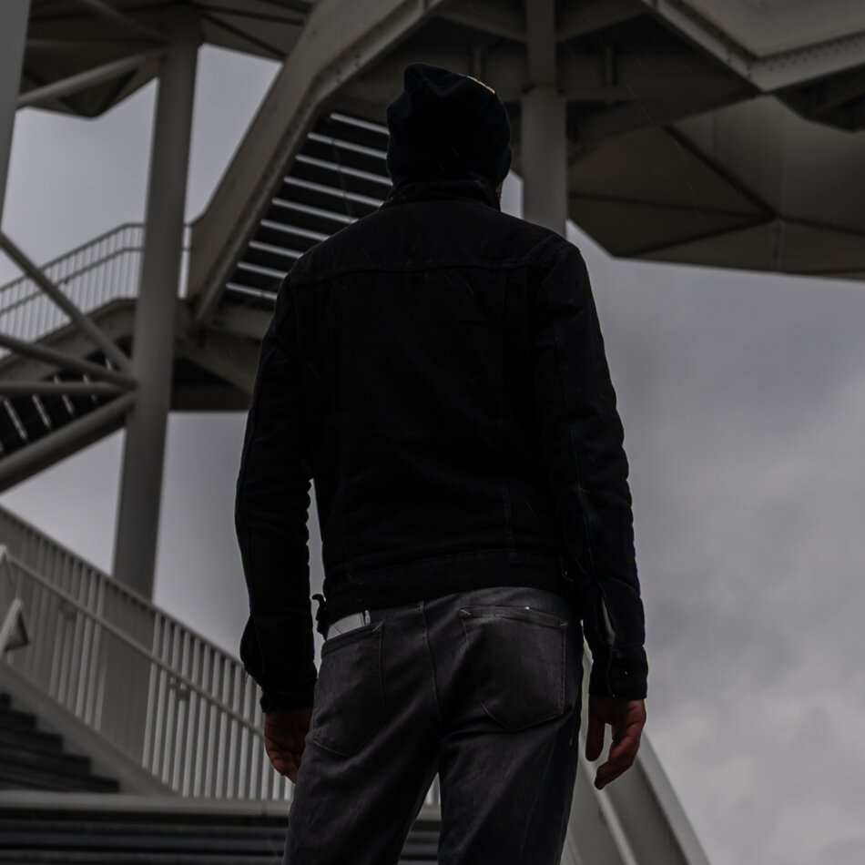 A monochromatic photo with hands in color. A man stands to the side wearing a deep black jacket, a deep black hat, and greyish/blackish trousers. In the background, there is a geometric metal structure primarily composed of stairs. The photo appears to be taken outdoors.