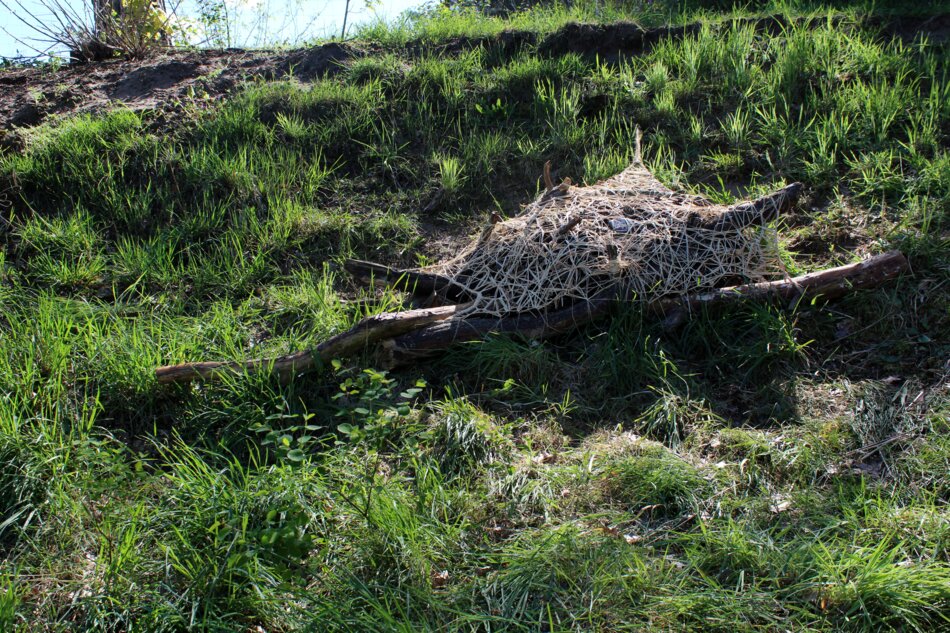 Artwork installation featuring natural materials such as branches and pinecones, combined with an artificial object, a gravestone fragment. Other materials include raffia, coir rope, and jute netting.