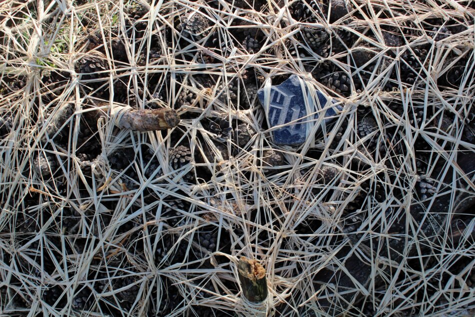 Mixed media artwork featuring pine cones, a gravestone fragment, and natural materials like raffia, coir rope, and jute netting.