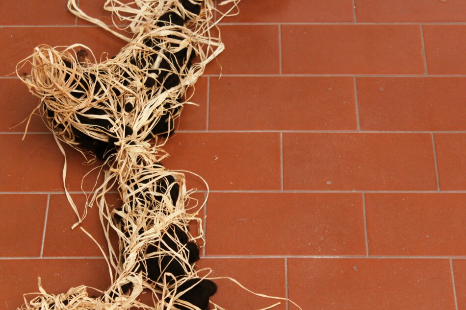 A close-up of raffia and black yarn tangled together, resembling a messy nest, on a red tiled floor.