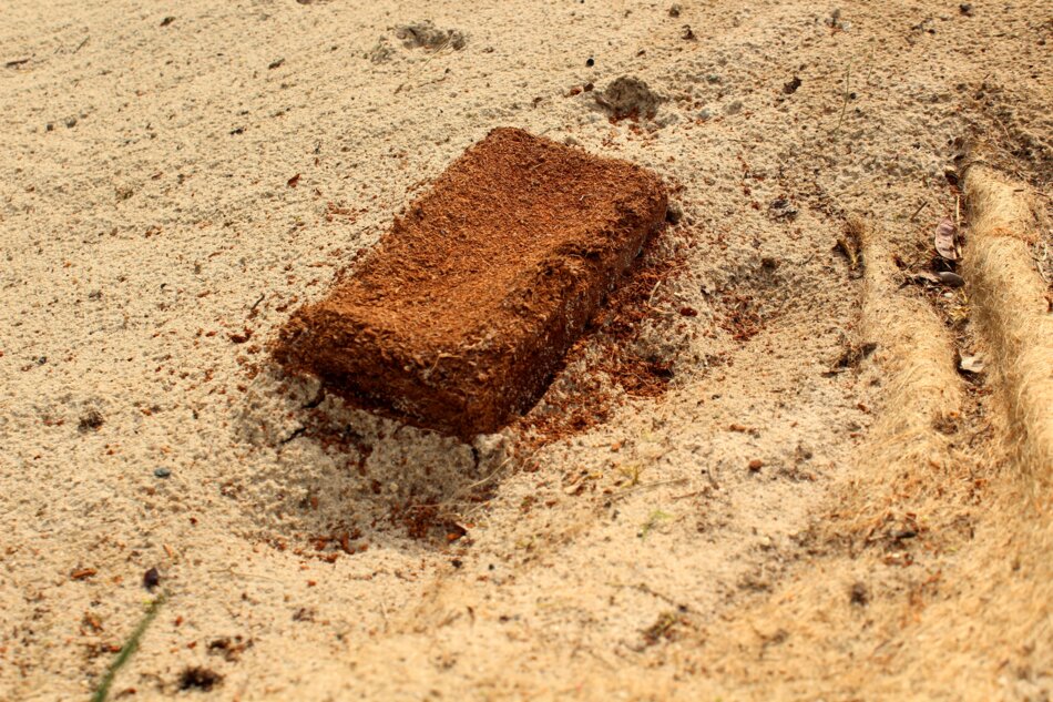 Contemporary art installation featuring a rectangular brown object resembling a brick on a sandy surface with fabric impressions.