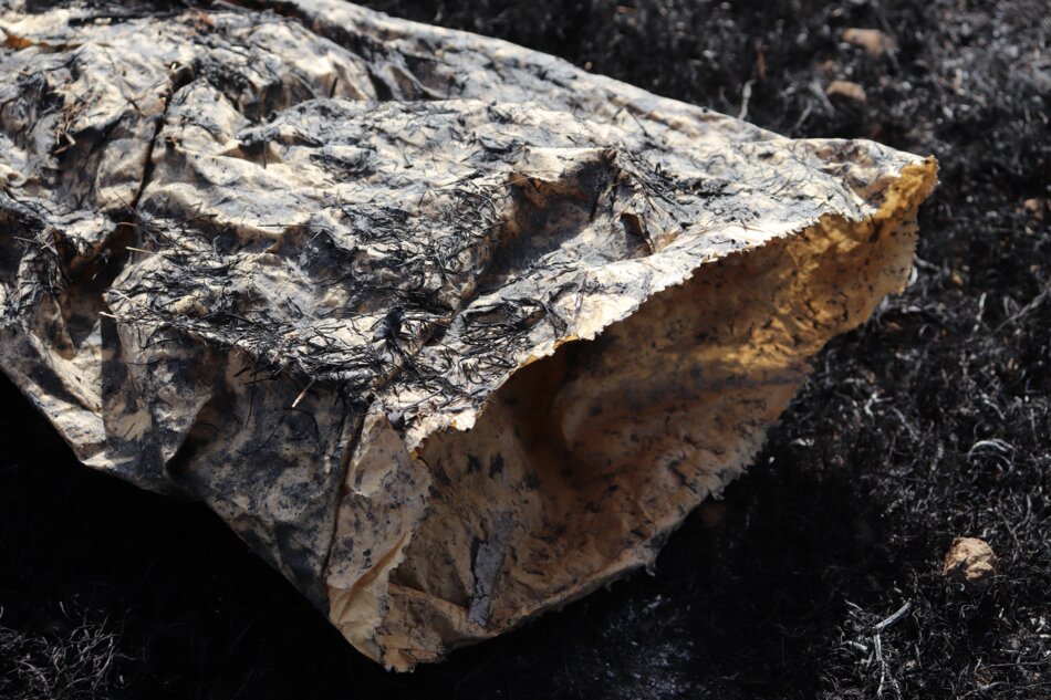 A charred paper bag lies on a patch of burned grass and soil. The bag is partially open, revealing its blackened interior.