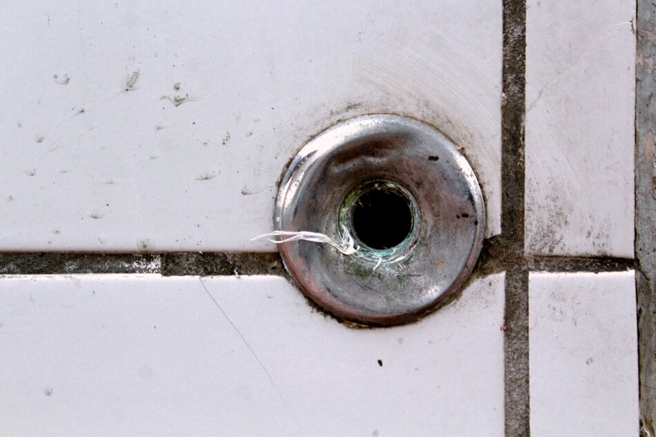 A close-up of a small, circular hole in a white tiled wall. The hole is surrounded by a metal ring.