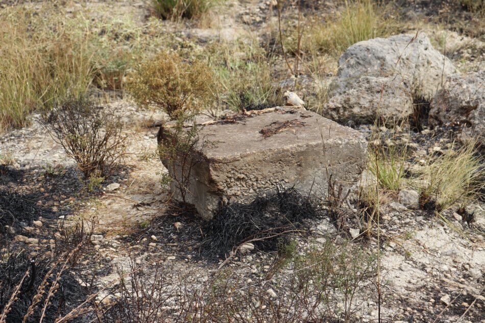 A weathered concrete block in a dry, grassy landscape with scattered rocks and burnt vegetation