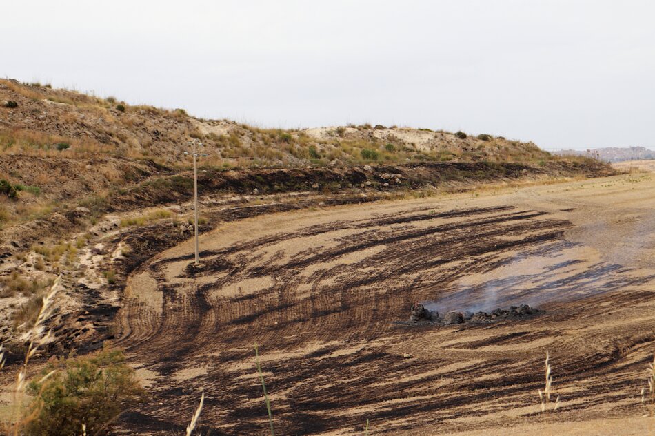 A wide shot of a scorched field with black, burned patches and smoke rising from a pile of smoldering debris. The field is surrounded by dry hills and a utility pole stands nearby.