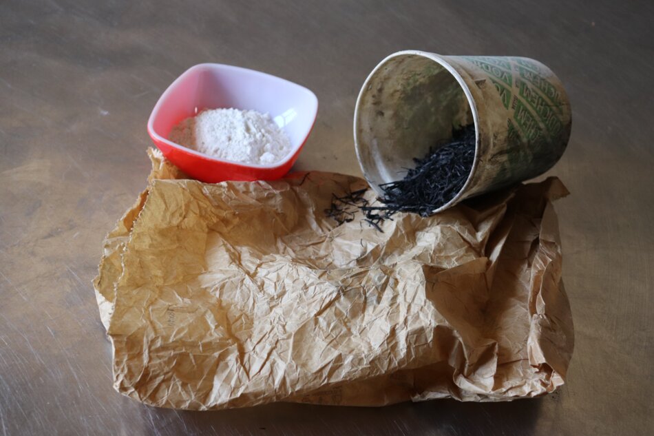 White powder in a red bowl and black powder in a white bucket, both placed on a brown paper bag.