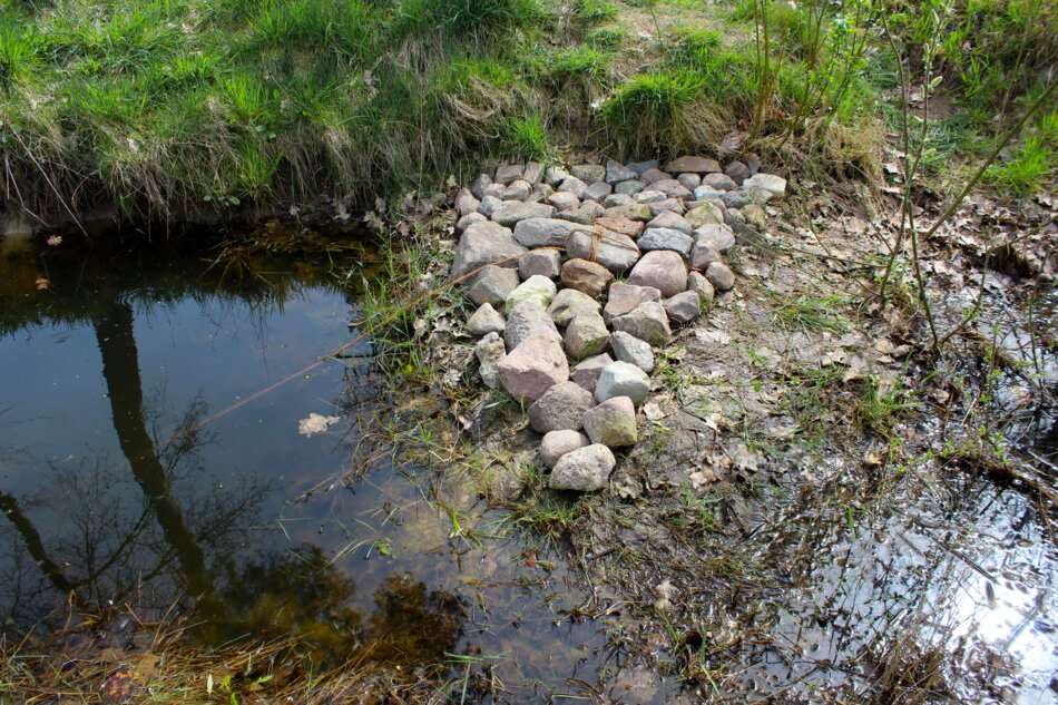 A pile of rocks forming a triangular shape at the edge of a pond. The pond is surrounded by green grass and a few trees.