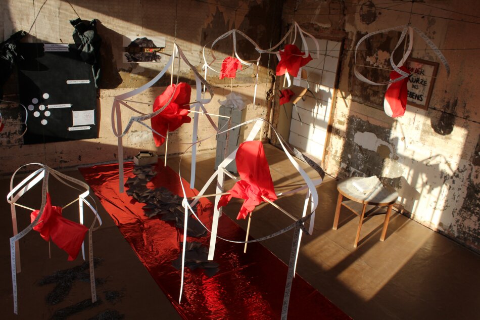 An installation art piece featuring a room filled with intricate structures made of white paper and draped with red fabric. The structures resemble architectural forms like arches and columns. Sunlight streams through windows, casting shadows on the walls and floor.