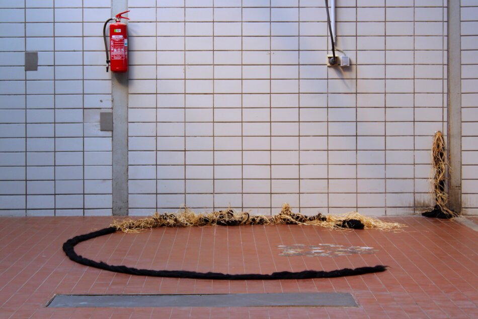 A large industrial hall with white tiled walls and a red fire extinguisher. A circular arrangement of chunky yarn lies on the red tiled floor.