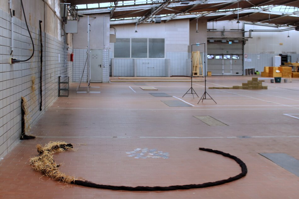 A large industrial space with white walls, a concrete floor, and a metal roof. A long, dark object resembling chunky yarn lies in a curved shape on the floor.