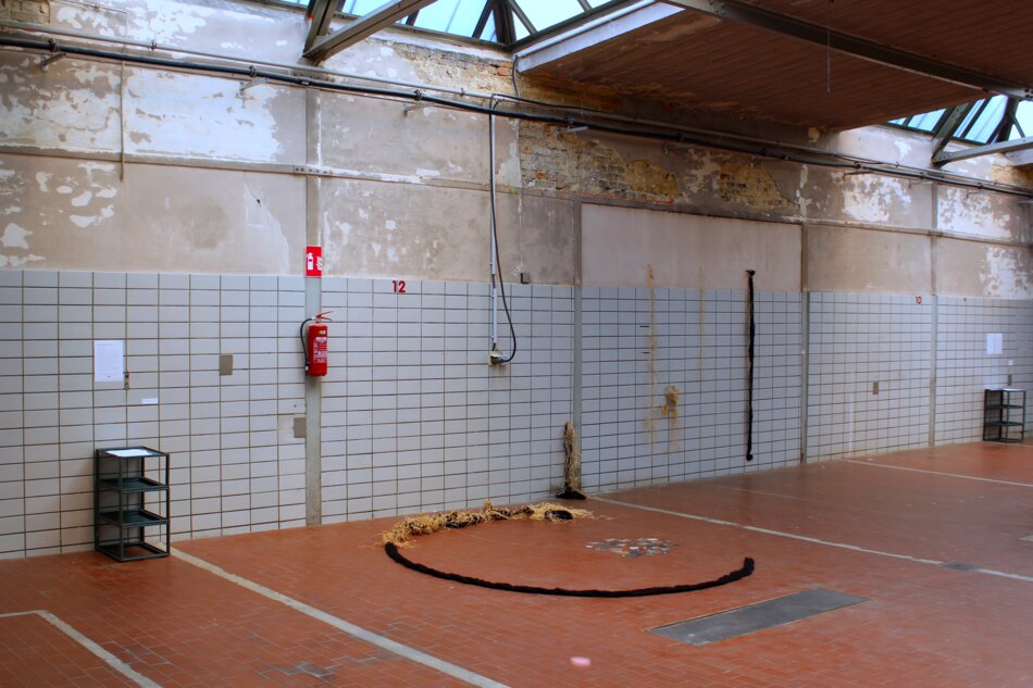 A large, abandoned industrial warehouse with white tiled walls, a concrete floor, and a skylight. A fire extinguisher hangs on the wall. A circular arrangement of chunky yarn lies on the floor, and various other art installations are visible in the background.