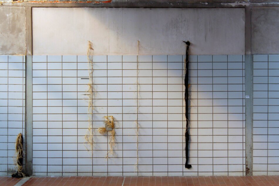 A wall with white tiles and a gray concrete beam above. Three vertical strings of twine hang down, one with a bundle of twigs attached. A long, dark object hangs down on the right.