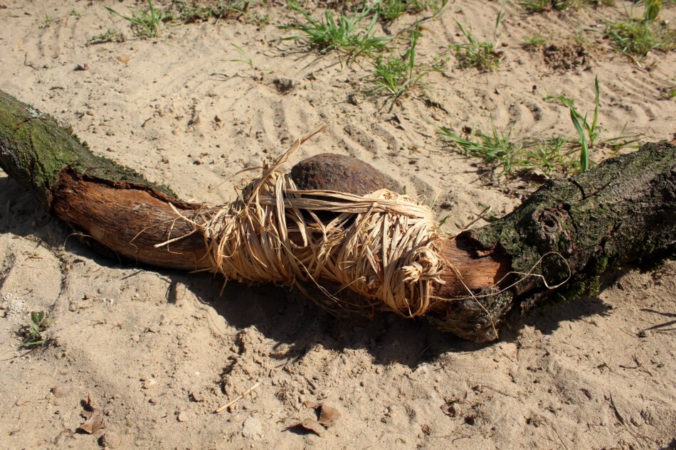 A close-up view of a metal object tied with raffia to a branch lying on sandy ground.