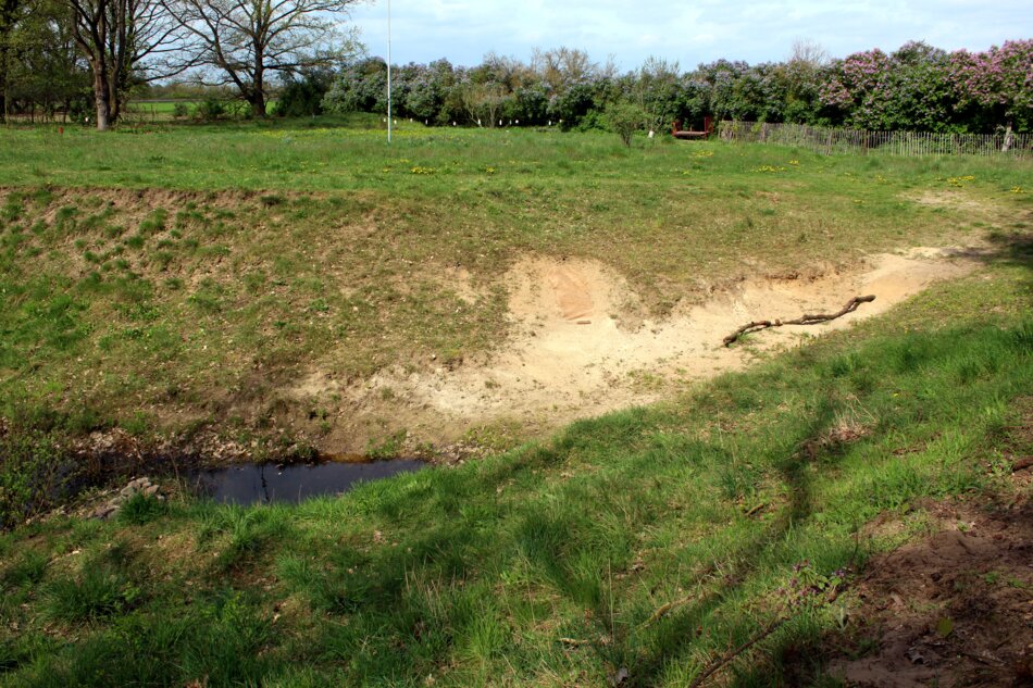 A shallow ditch with exposed soil and a small stream running through it. The ditch is surrounded by grassy fields and a few trees in the background.
