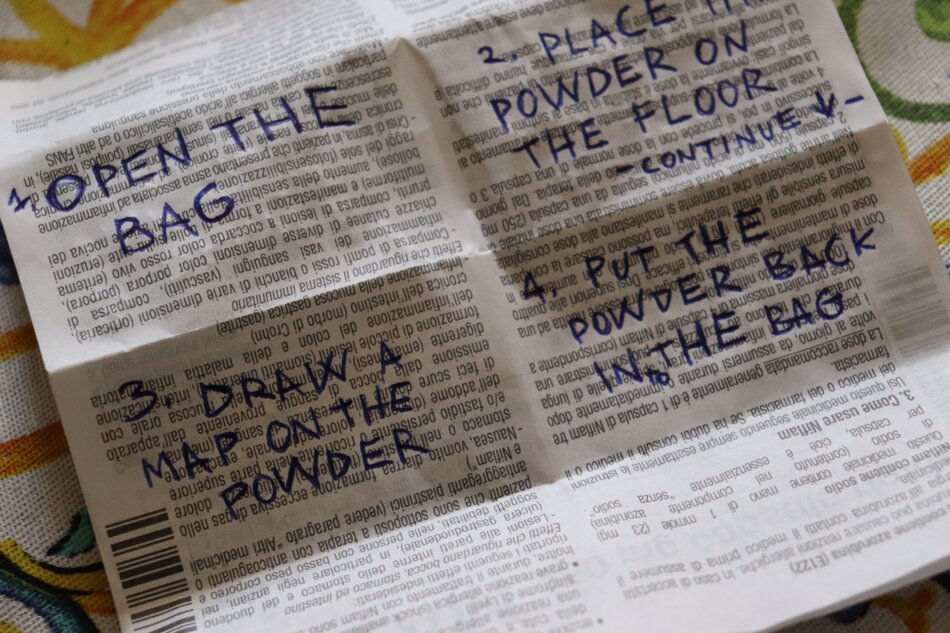 A close-up of a folded paper with handwritten instructions in blue ink. The instructions are to open the bag, place the powder on the floor, draw a map on the powder, and put the powder back in the bag.