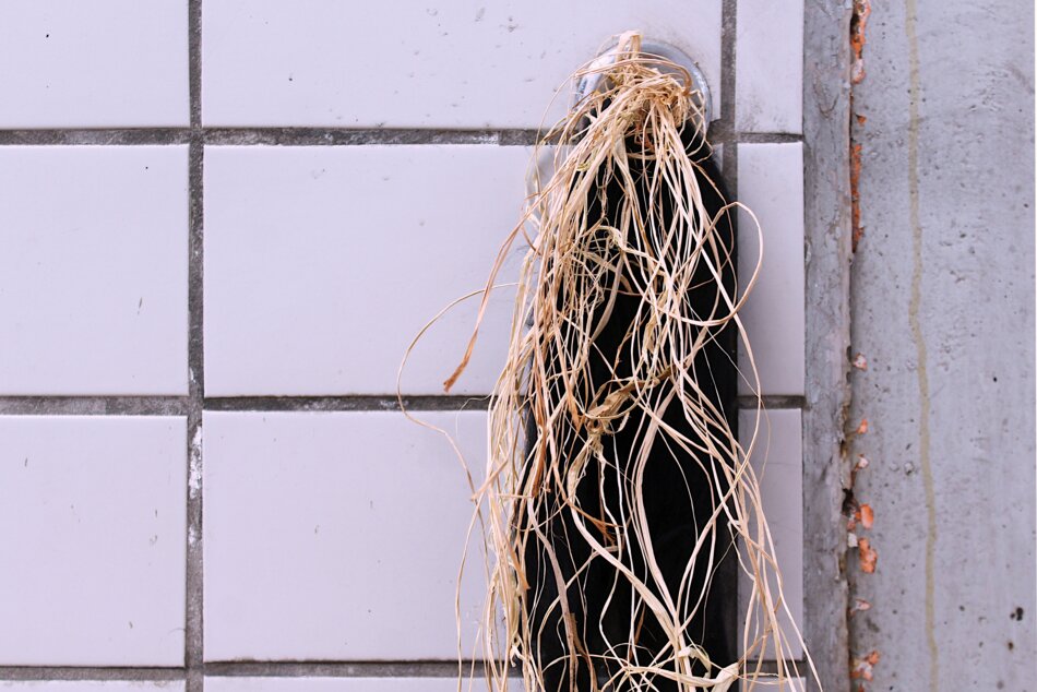 A bundle of brown raffia hanging from a circular hole in a white tiled wall. The hole is surrounded by a metal ring.