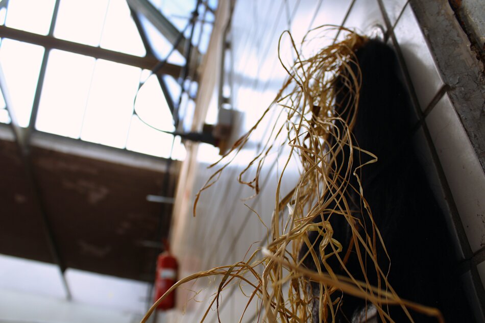 A bundle of brown raffia hanging from a black object mounted on a white tiled wall. A fire extinguisher and a light fixture are visible in the background.