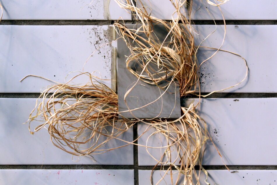 A bundle of brown raffia surrounding a gray object mounted on a white tiled wall.