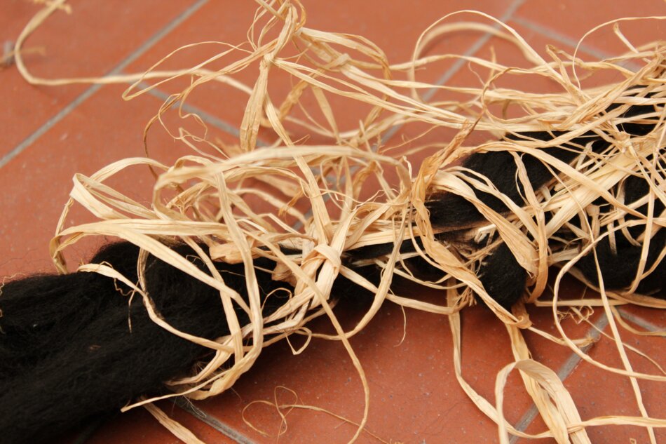 A close-up of raffia and black yarn intertwined, resembling a tangled nest, on a red tiled floor.