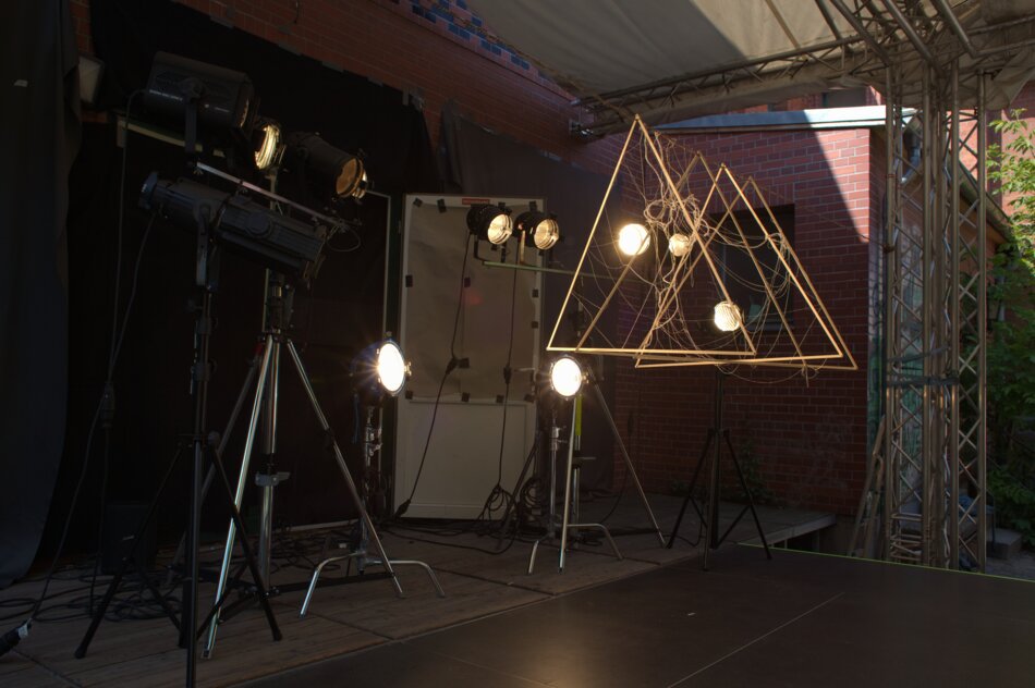 Exterior view of a human-scale camera obscura structure (not accessible to viewers) made of four linden wood strip triangles, seaweed threads, and polypropylene fibers, with nine theatrical lights mounted on metal supports on an outdoor stage.