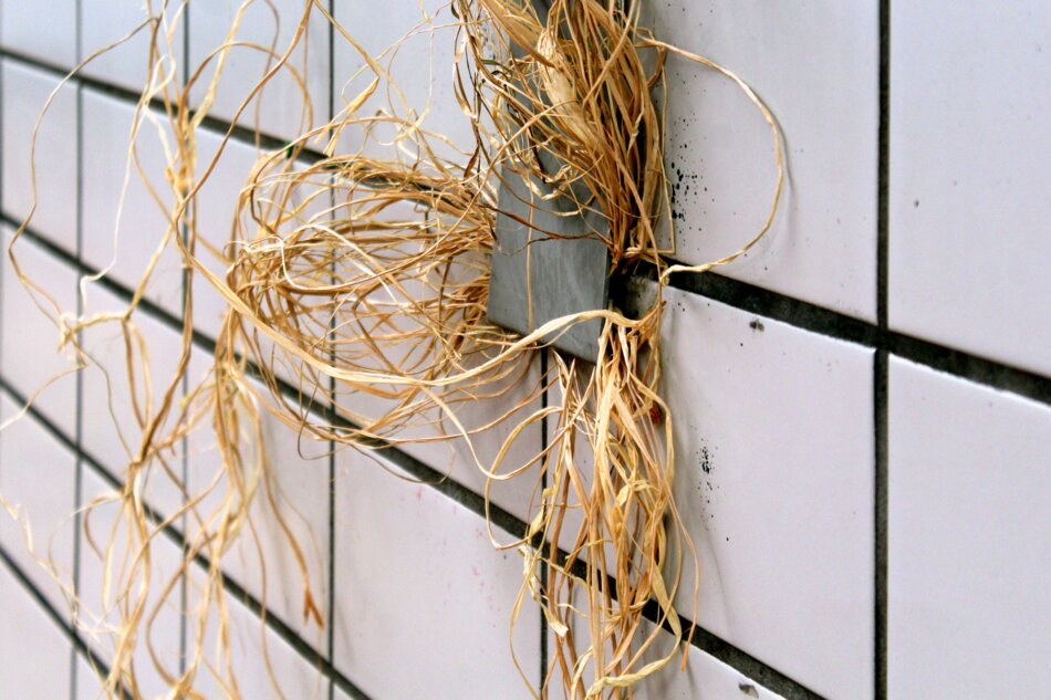 A bundle of brown raffia surrounding a gray object mounted on a white tiled wall.