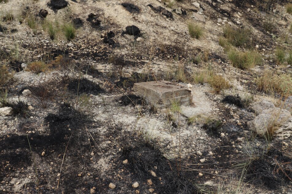 A concrete block stands on a fire-damaged hillside. The surrounding ground is covered in black ash and remnants of burned vegetation.