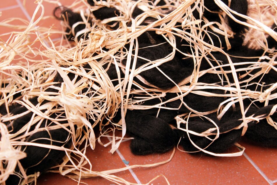 A close-up of raffia and black yarn tangled together, resembling a messy nest, on a red tiled floor.