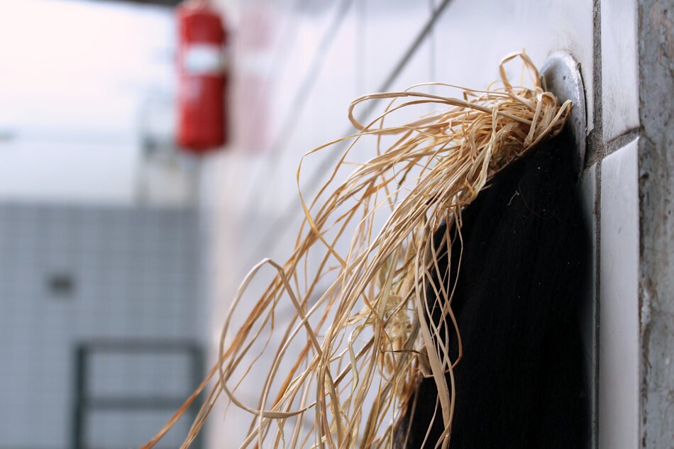 A bundle of brown raffia hanging from a black object mounted on a white tiled wall. A red fire extinguisher is visible in the background.