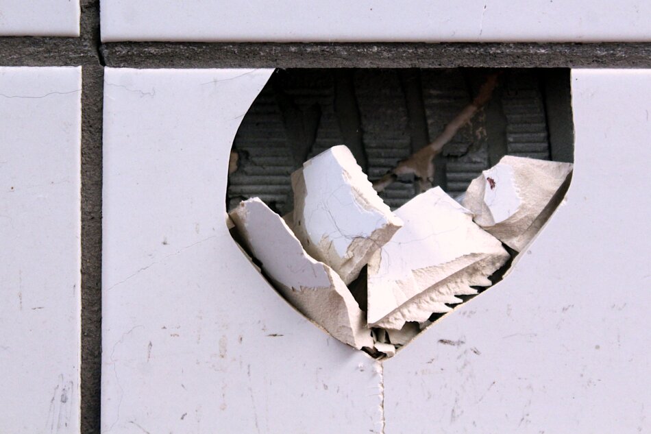 A close-up of a large, rectangular hole in a white tiled wall. The hole has jagged edges and contains broken pieces of tile.