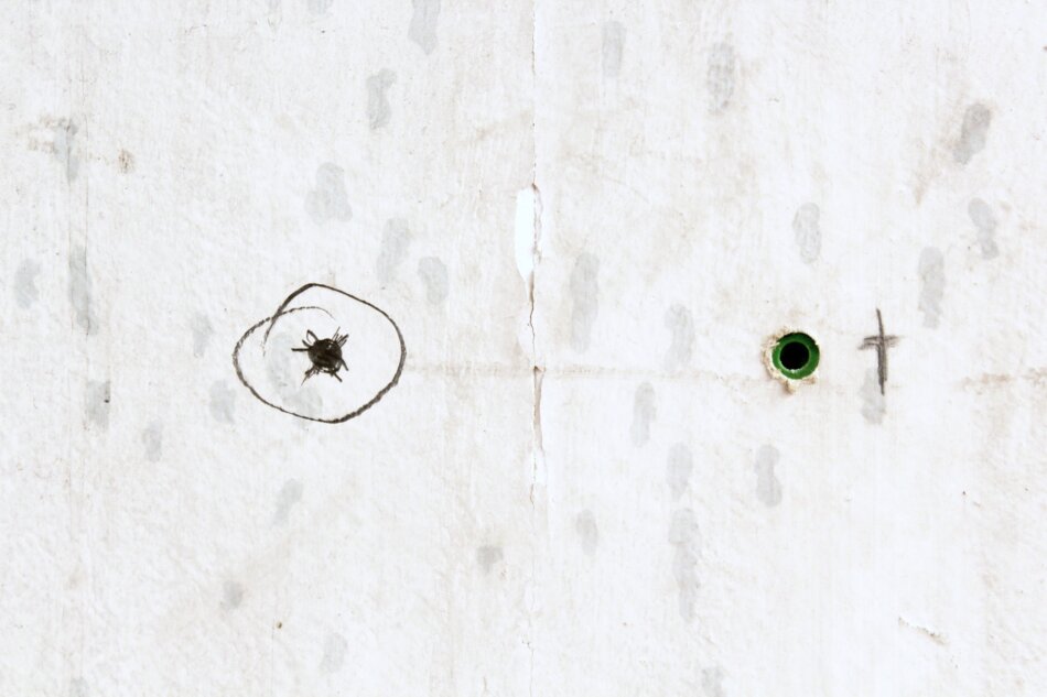 A close-up of a white wall with two marks on it. One mark is a circular bullet hole with a surrounding ring, and the other is a green cross.